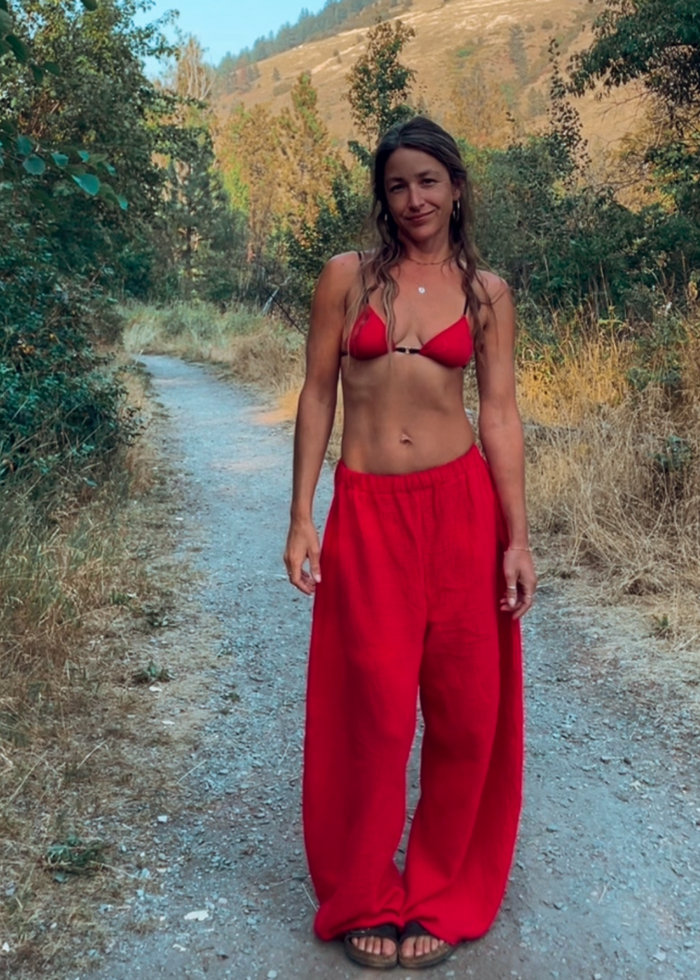 A woman stands on a dirt path outdoors wearing a bright red two-piece swimsuit with a triangle top and very wide-leg pants, with trees and a hill in the background.